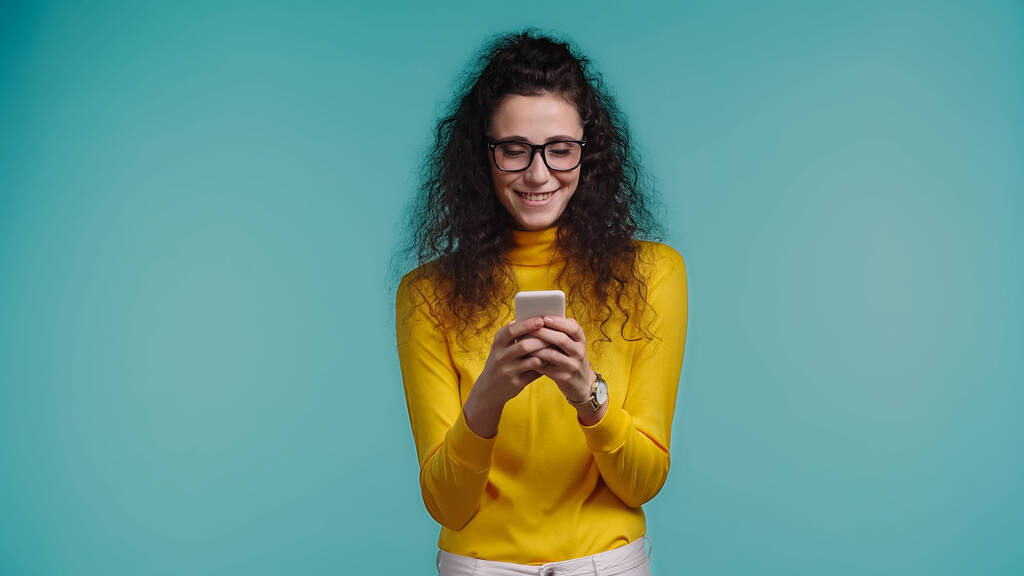 радостная молодая женщина в очках смс на смартфоне изолированы на синий  - Фото, изображение