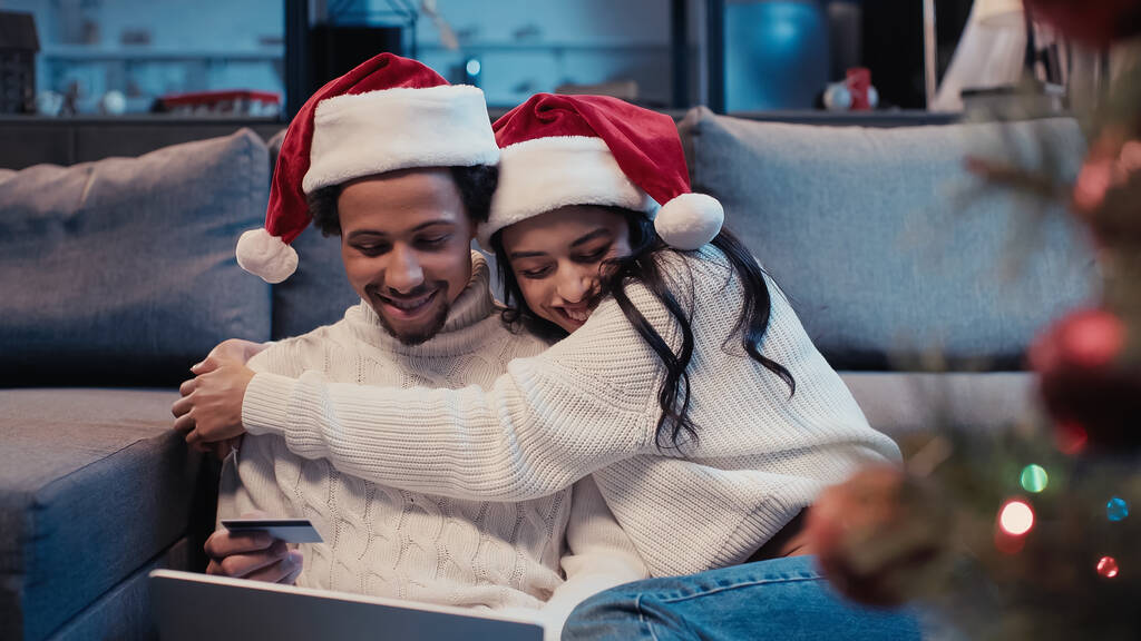 heureux couple afro-américain dans santa chapeaux câlins tandis que les achats en ligne près flou arbre de Noël  - Photo, image