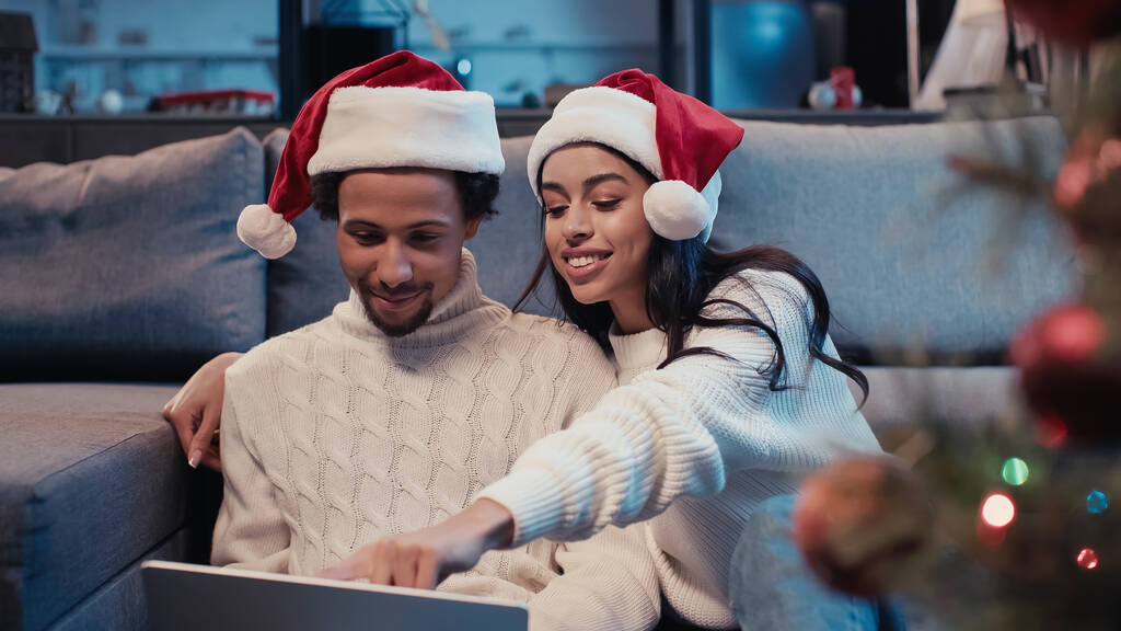 heureux couple afro-américain chapeaux santa en utilisant un ordinateur portable près de l'arbre de Noël flou  - Photo, image