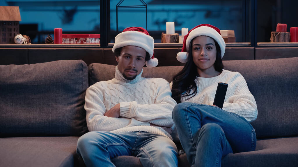 sorridente donna afro-americana guardando film con uomo annoiato in cappello di Babbo Natale - Foto, immagini
