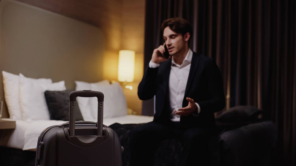 jovem empresário desfocado falando no smartphone perto da mala no quarto de hotel - Foto, Imagem