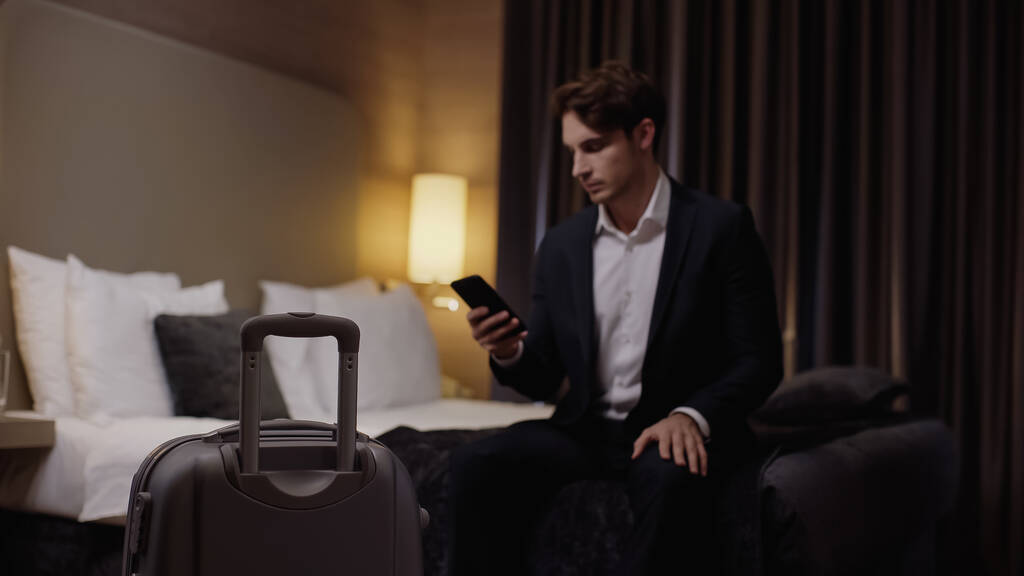 rozmazaný mladý podnikatel držící smartphone v blízkosti kufru v hotelovém pokoji - Fotografie, Obrázek