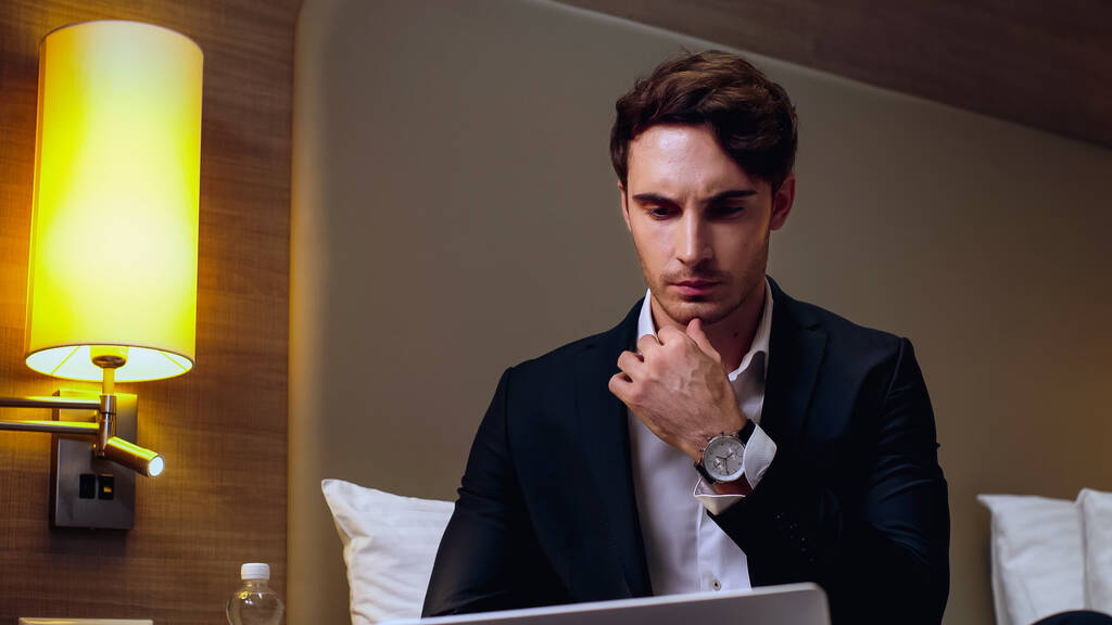 στοχαστικός νεαρός επιχειρηματίας που χρησιμοποιεί φορητό υπολογιστή στο δωμάτιο του ξενοδοχείου - Φωτογραφία, εικόνα