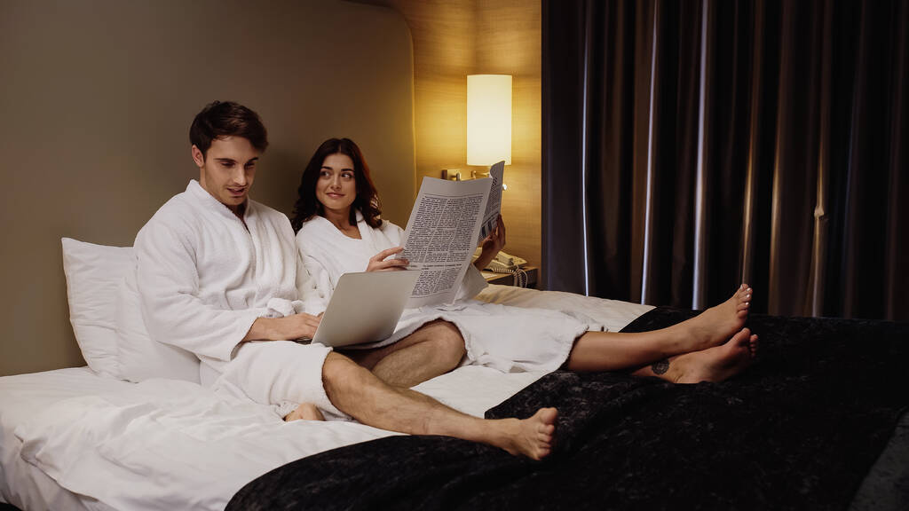 man using laptop near cheerful girlfriend with newspaper resting in hotel room - Photo, Image