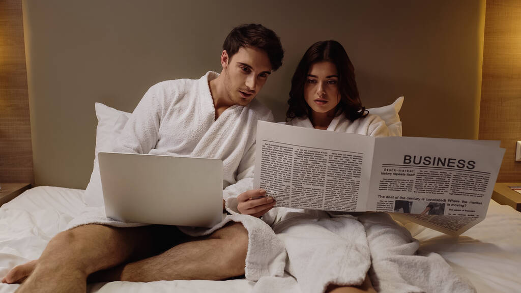 ホテルの部屋にいるガールフレンドの近くでノートパソコンを読むビジネス新聞の男 - 写真・画像