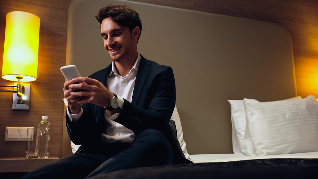 gelukkig zakenman zitten op bed en chatten op de smartphone in de hotelkamer - Foto, afbeelding