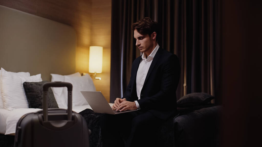 mladý seriózní podnikatel sedí na posteli a pomocí notebooku v blízkosti zavazadel v hotelovém pokoji  - Fotografie, Obrázek