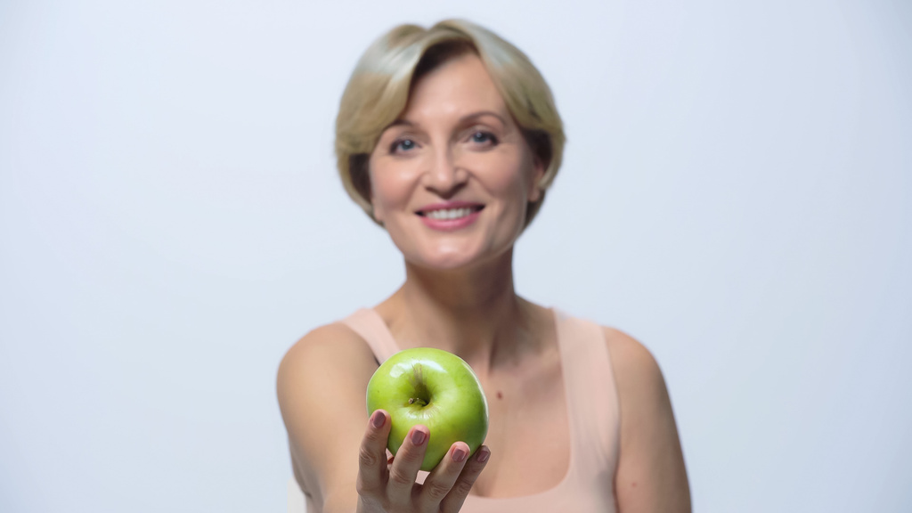 femme d'âge moyen floue montrant pomme verte et souriant à la caméra isolé sur blanc - Photo, image