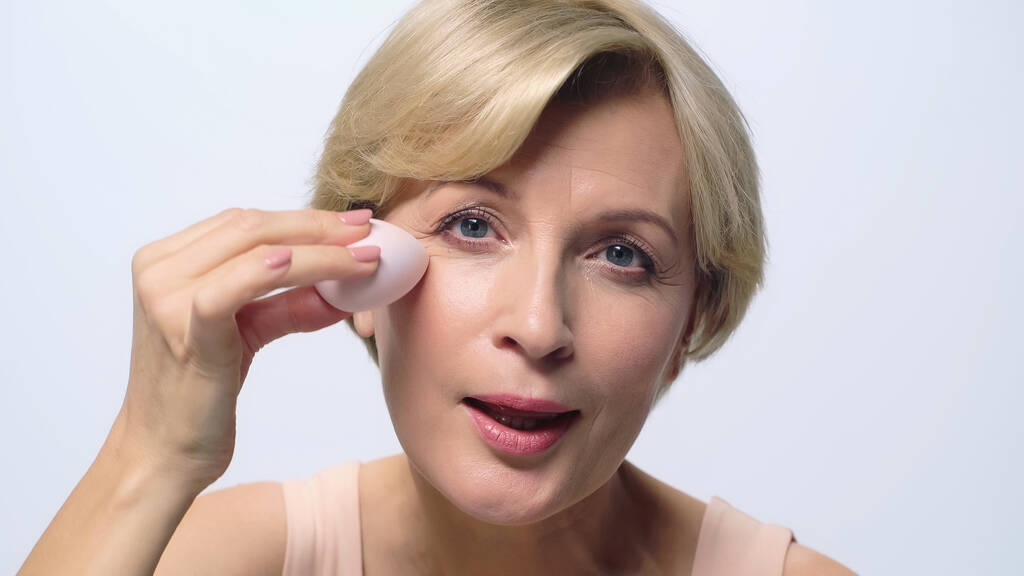 Frau mittleren Alters blickt in die Kamera, während sie Gesicht Foundation mit kosmetischen Puff isoliert auf weiß aufträgt - Foto, Bild