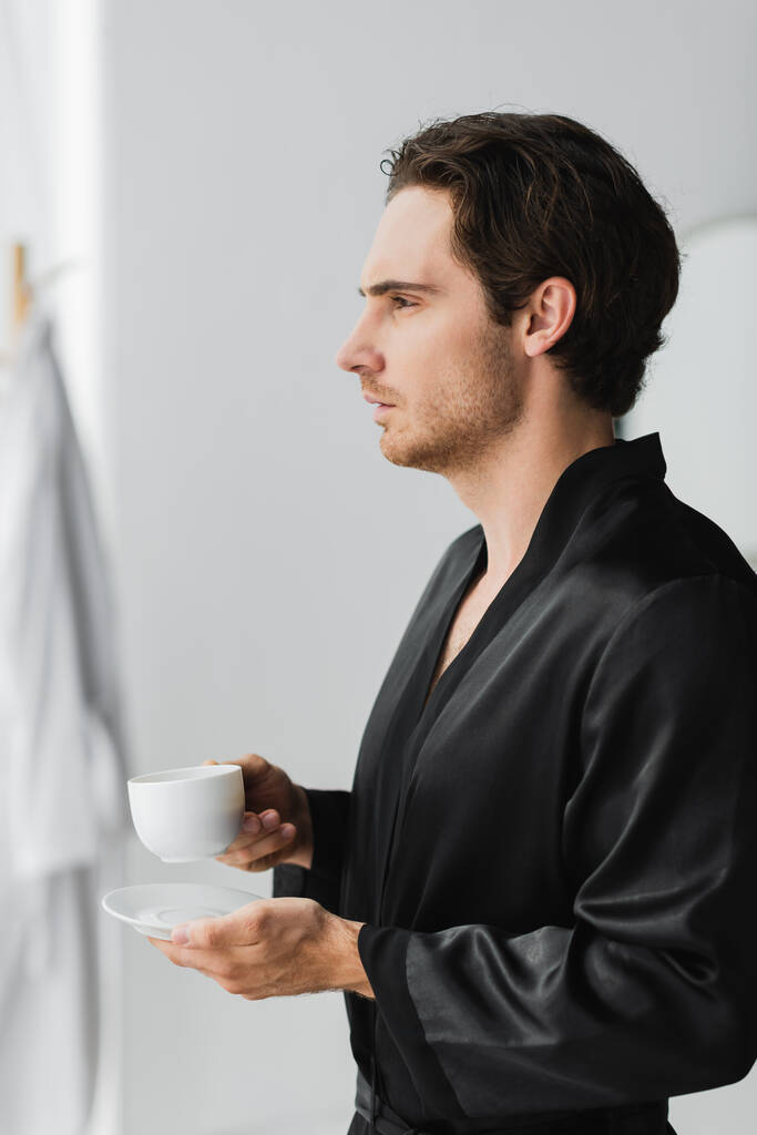 Vista lateral do homem em robe de seda segurando xícara de café no banheiro  - Foto, Imagem