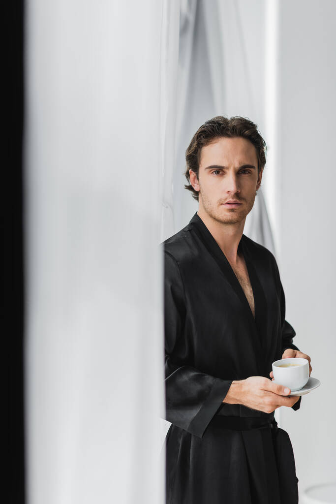 Jonge man in zijden badjas met koffie in de buurt van gordijnen thuis  - Foto, afbeelding