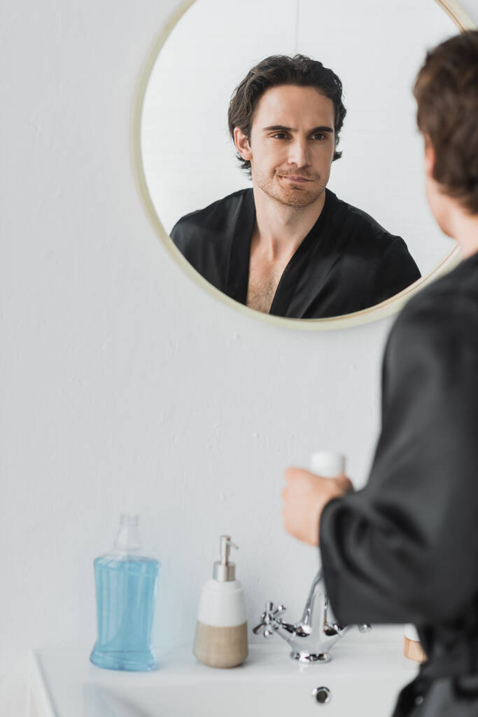 Homme flou en robe de soie rinçant la bouche près du miroir à la maison  - Photo, image