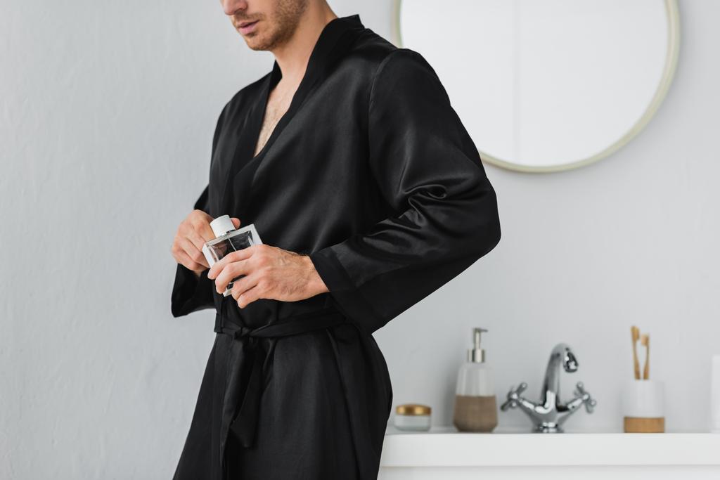 Vista cortada do homem em robe de cetim segurando perfume no banheiro  - Foto, Imagem