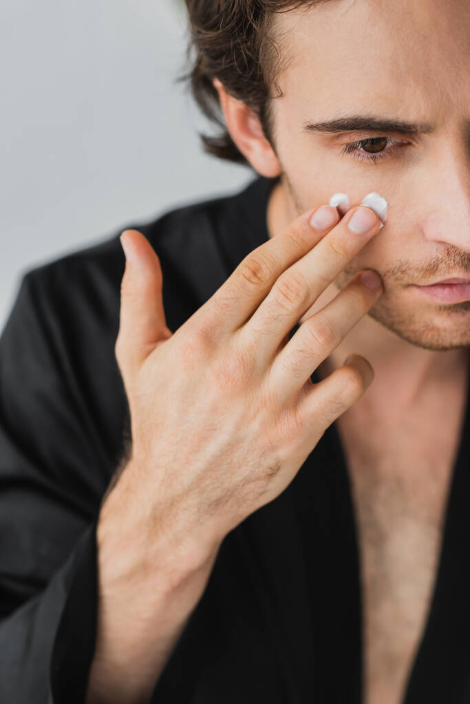 Cropped view of young man in black robe applying cosmetic cream on face isolated on grey  - Photo, Image