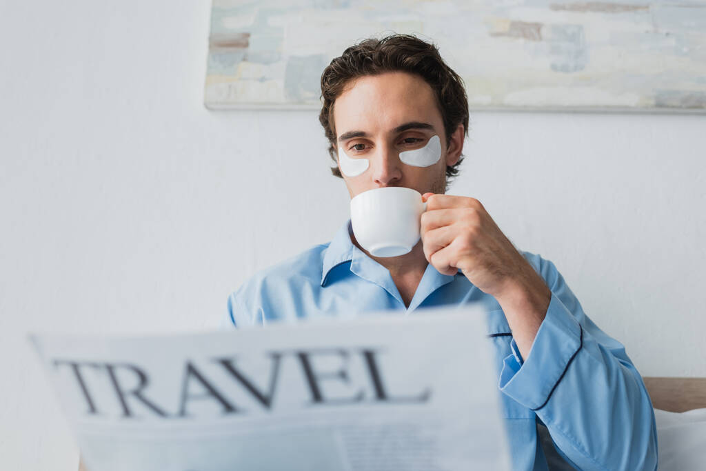 Fiatal férfi szemkötőben és pizsamában kávét iszik és homályos újságot olvas a hálószobában.  - Fotó, kép