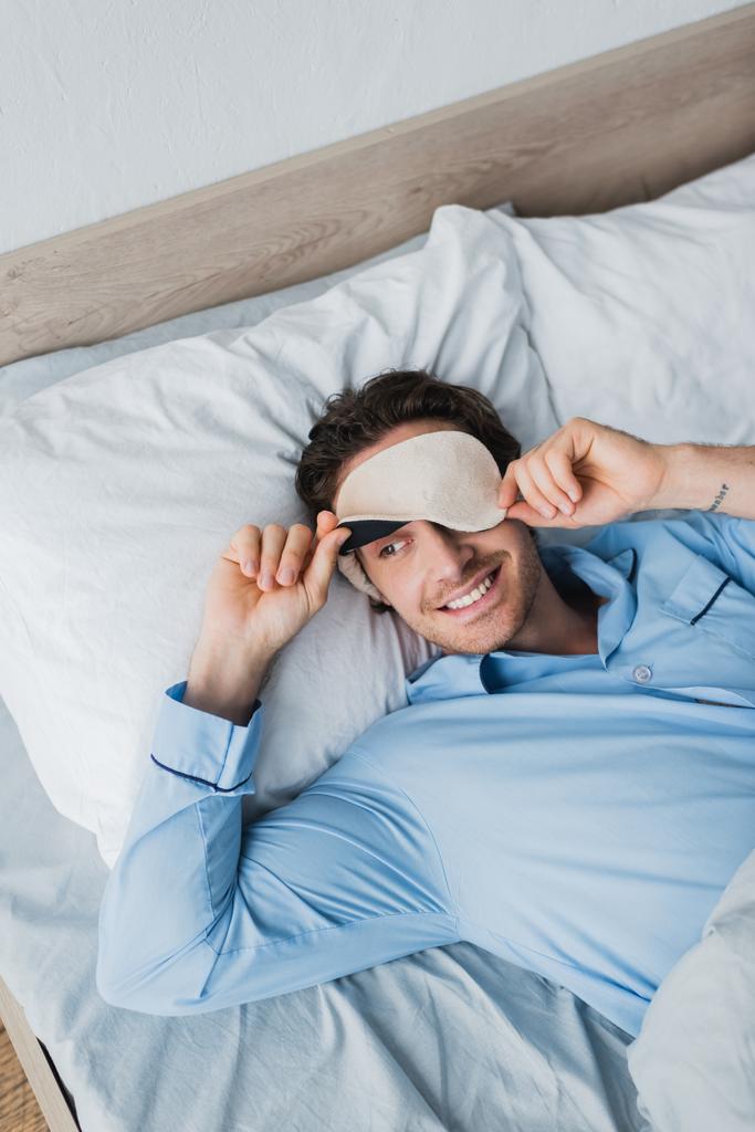 Yüksek açılı, uyku maskeli ve pijamalı gülümseyen adamı sabah yatakta yatarken görüyorum.  - Fotoğraf, Görsel