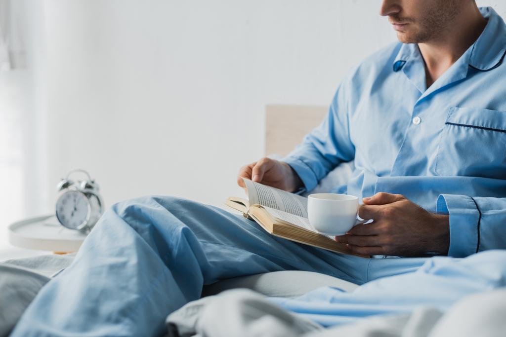 Vista ritagliata dell'uomo in pigiama che tiene la tazza e legge il libro vicino alla sveglia offuscata in camera da letto  - Foto, immagini