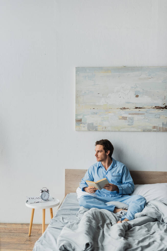 Young man in pajama holding book near alarm clock on bed in morning  - Photo, Image