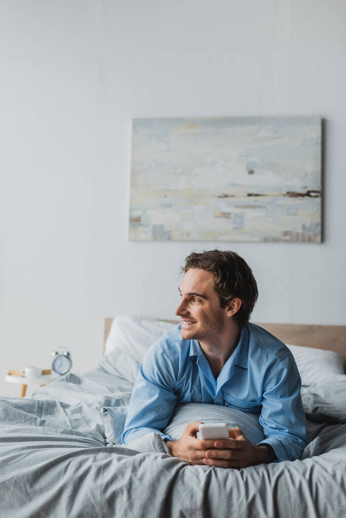 Uomo sorridente in pigiama utilizzando il cellulare mentre sdraiato sul letto al mattino  - Foto, immagini