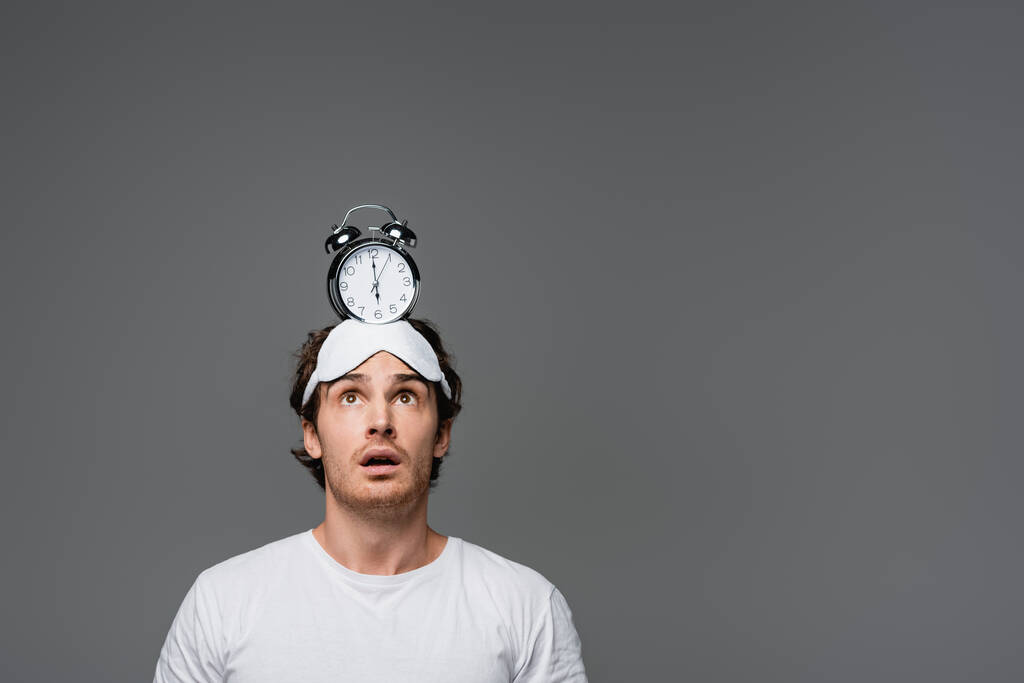 Schockierter Mann mit Schlafmaske blickt auf Wecker am Kopf isoliert auf grau  - Foto, Bild