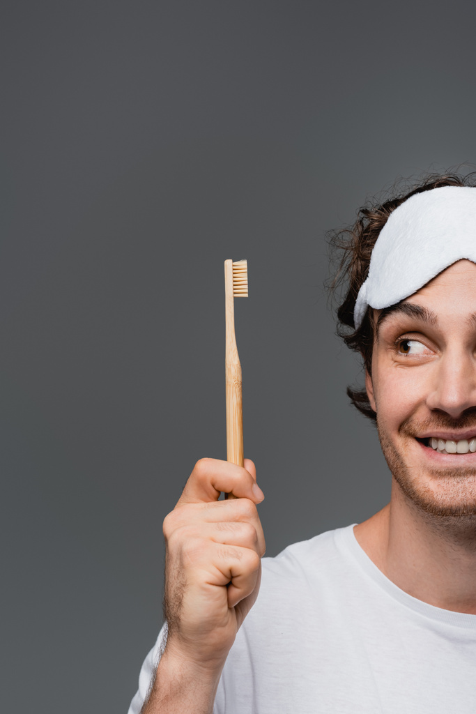 Cropped view of smiling man in sleep mask looking at toothbrush isolated on grey  - Photo, Image