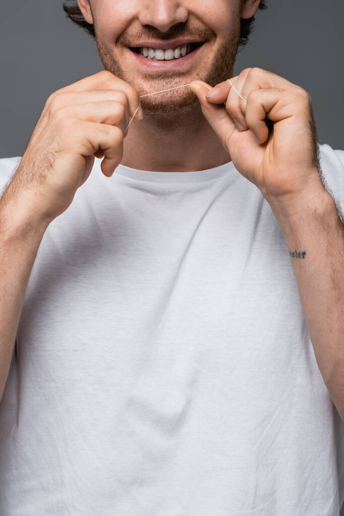 Vista recortada del hombre positivo en camiseta blanca sosteniendo hilo dental aislado en gris  - Foto, Imagen