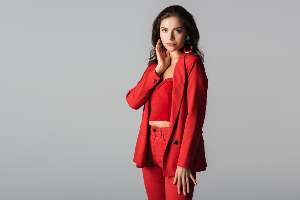 junge stylische Frau in rotem Anzug und Ohrringen posiert isoliert auf grau - Foto, Bild