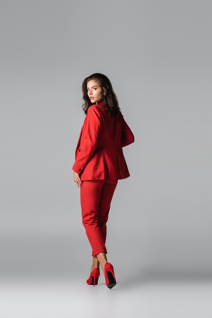 повна довжина молодої стильної жінки на червоних підборах і костюм позує під час ходьби на сірому
 - Фото, зображення
