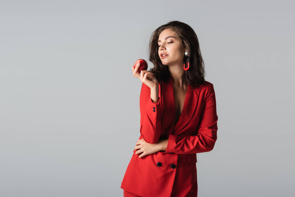 stylowa kobieta w czerwonym garniturze pozowała ze świeżym jabłkiem odizolowanym na szaro  - Zdjęcie, obraz