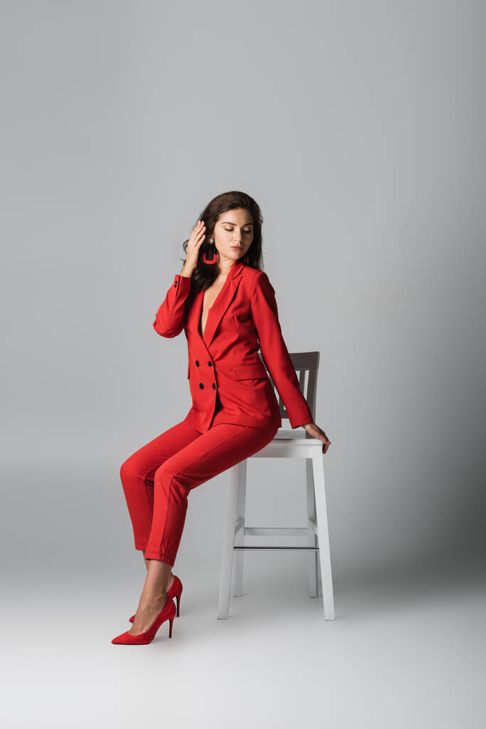полная длина модной женщины в красном костюме, позирующей сидя на белом стуле на сером - Фото, изображение