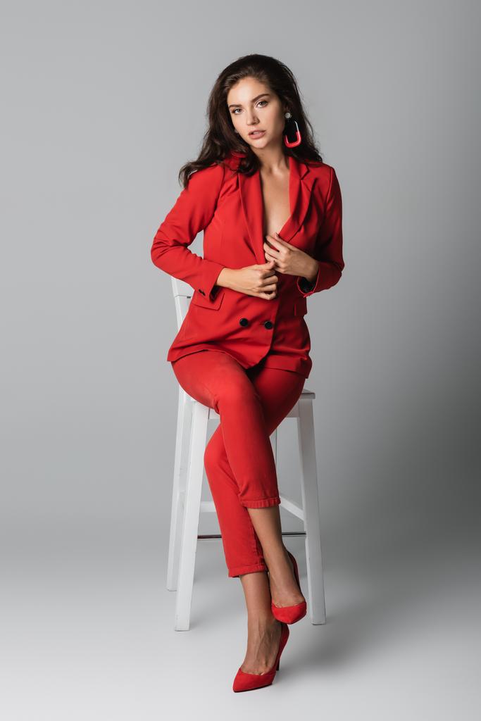 pleine longueur de jeune femme sexy en costume rouge posant tout en étant assis sur une chaise blanche sur gris - Photo, image
