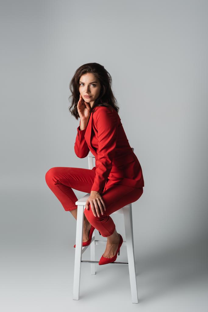 женщина в красном костюме в полный рост, сидящая на белом стуле на сером - Фото, изображение