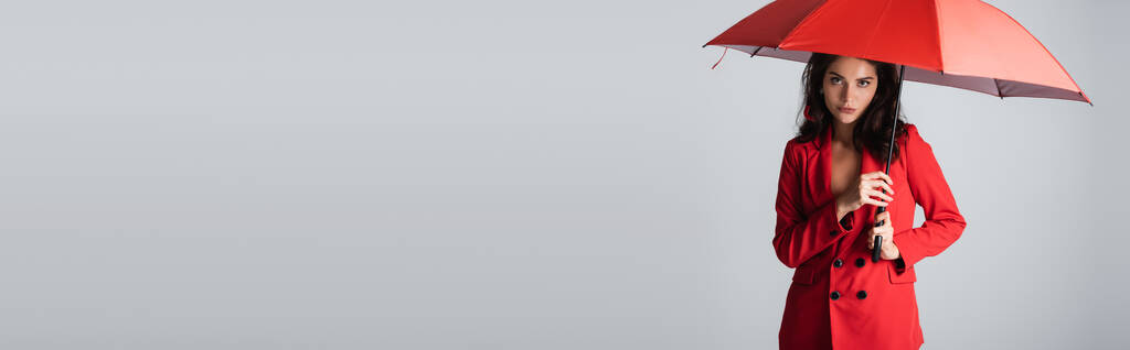 молодая брюнетка в красном костюме стоящая под зонтиком изолированная на сером баннере - Фото, изображение