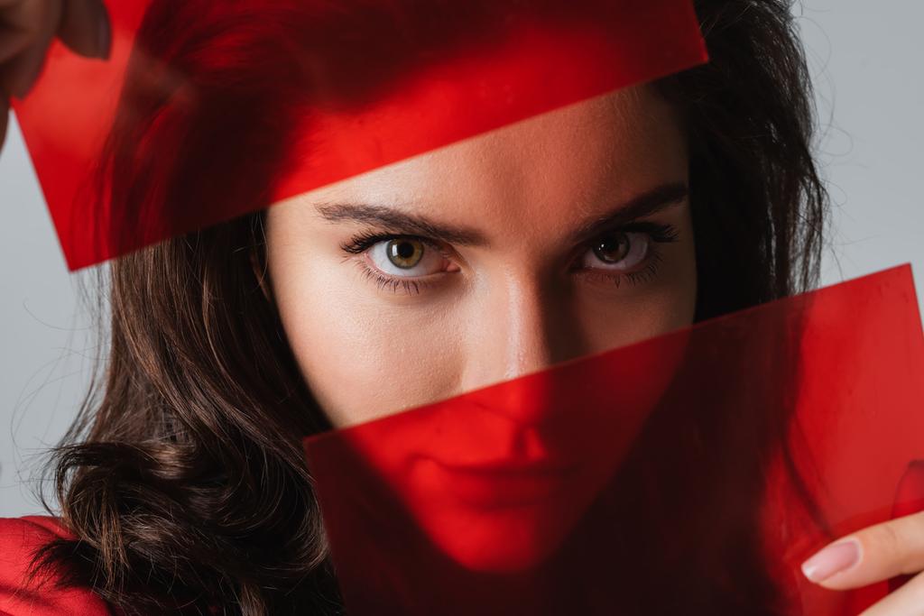 fiatal nő néz kamera keresztül piros üveg elszigetelt szürke - Fotó, kép
