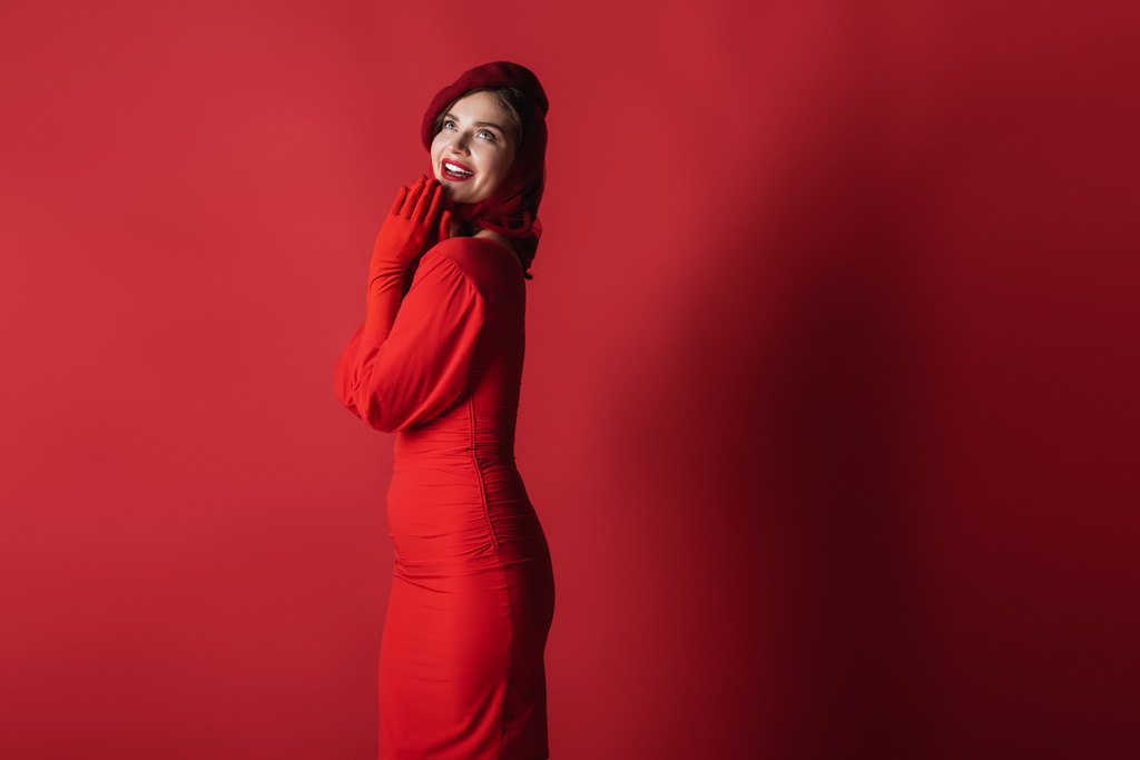 fröhliche und elegante Frau in Baskenmütze und Kleid posiert auf Rot - Foto, Bild