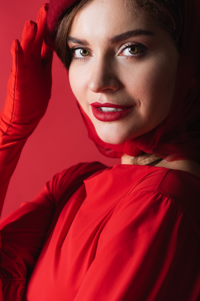 veselá mladá žena v rukavici upravující baret izolované na červené - Fotografie, Obrázek