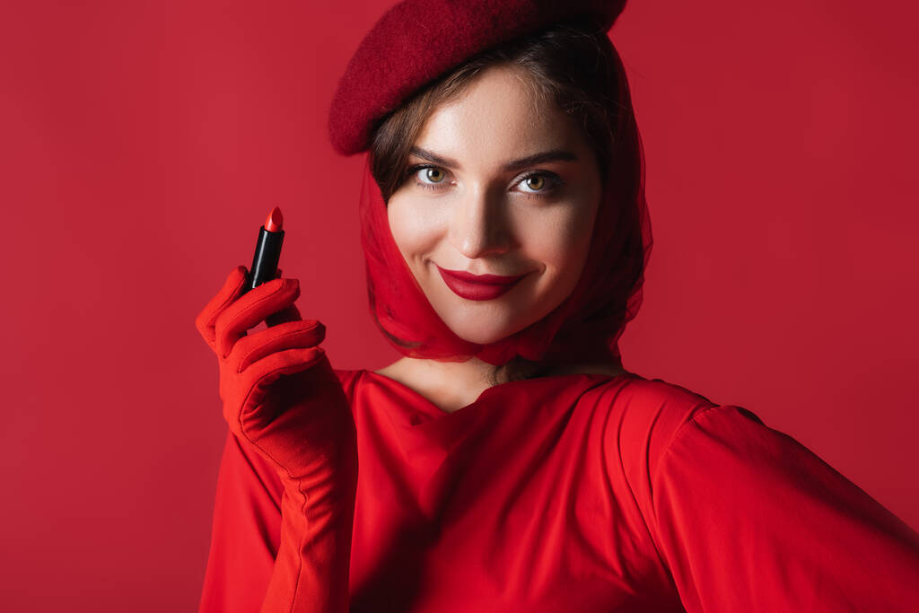 happy woman in glove, headscarf and beret holding lipstick isolated on red - Photo, Image