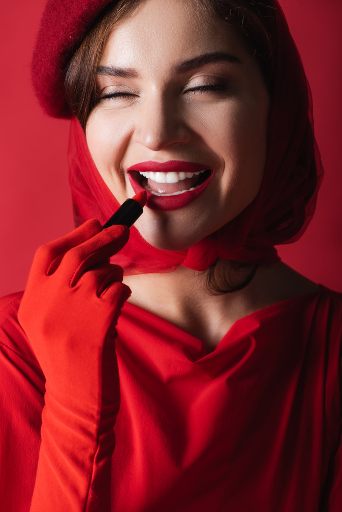 mulher alegre em luva, lenço de cabeça e boina aplicando batom isolado no vermelho - Foto, Imagem