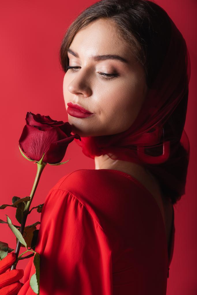 pretty young woman in glove and headscarf holding rose isolated on red - Photo, Image