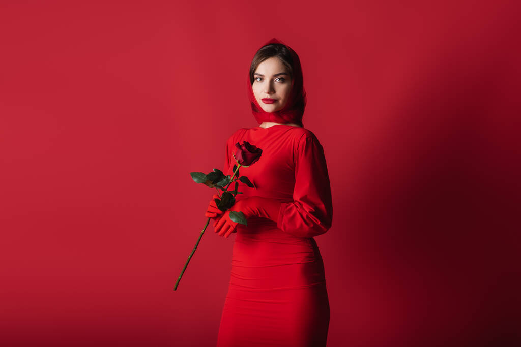 elegante jonge vrouw in jurk, handschoenen en hoofddoek holding roos op rood - Foto, afbeelding