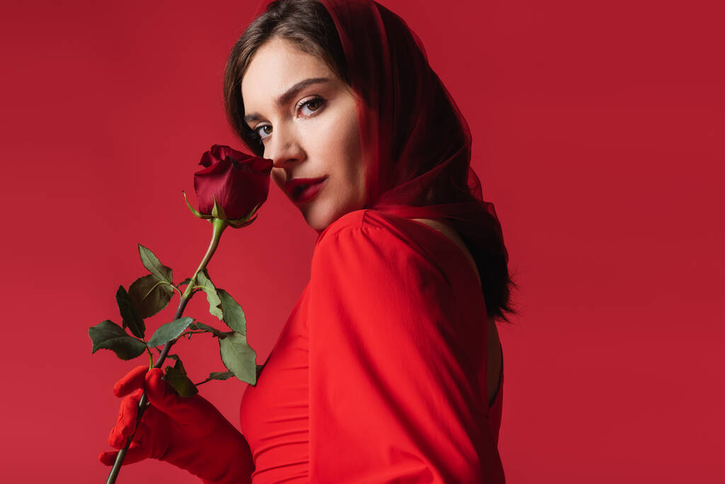 young woman in dress and headscarf smelling rose while looking at camera isolated on red - Photo, Image