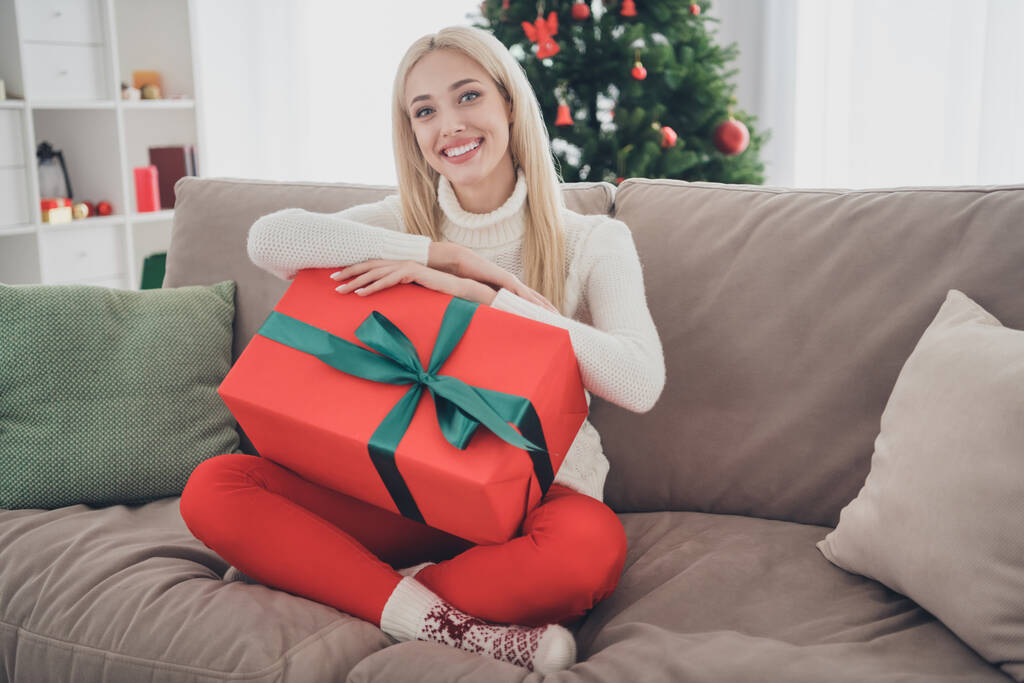 Полноразмерное фото женщины в белом пуловере, которая держит большую подарочную коробку, сидя дома на диване - Фото, изображение