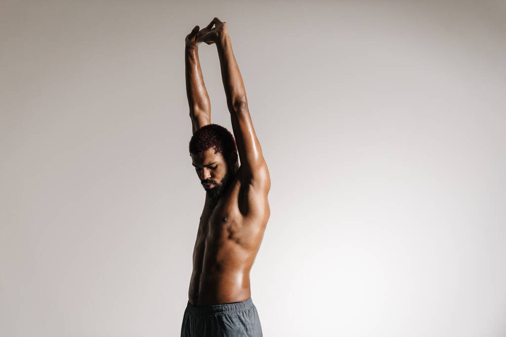 Schwarzer Sportler ohne Hemd beim Stretching, während er isoliert vor weißem Hintergrund trainiert - Foto, Bild