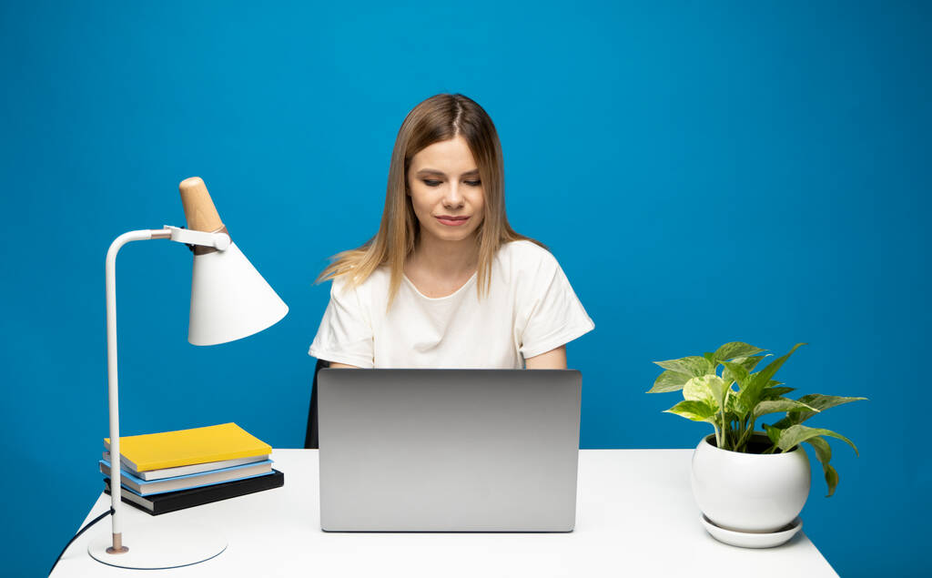 Portret van een mooie jonge vrouw die studeert terwijl ze aan tafel zit met een grijze laptop, notebook. glimlachende zakenvrouw werken met een laptop geïsoleerd op een blauwe achtergrond. - Foto, afbeelding