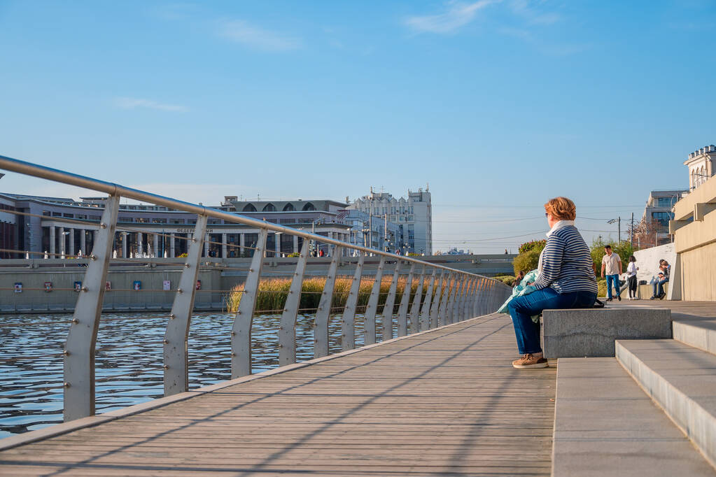 Kazan, Russie - 14 octobre 2020 : Le remblai du lac de la ville avec un sentier piétonnier en bois et une clôture métallique. Une femme est assise seule sur un banc près de l'eau au coucher du soleil. - Photo, image