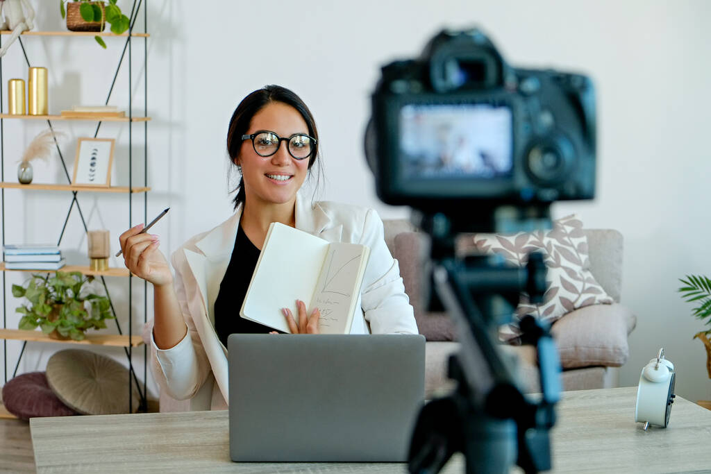 Young smart looking woman wearing glasses recording a video lesson on a camera. Portrait of female teacher by the desk with a textbook explaining a subject. Close up, copy space, brick wall background - Photo, Image