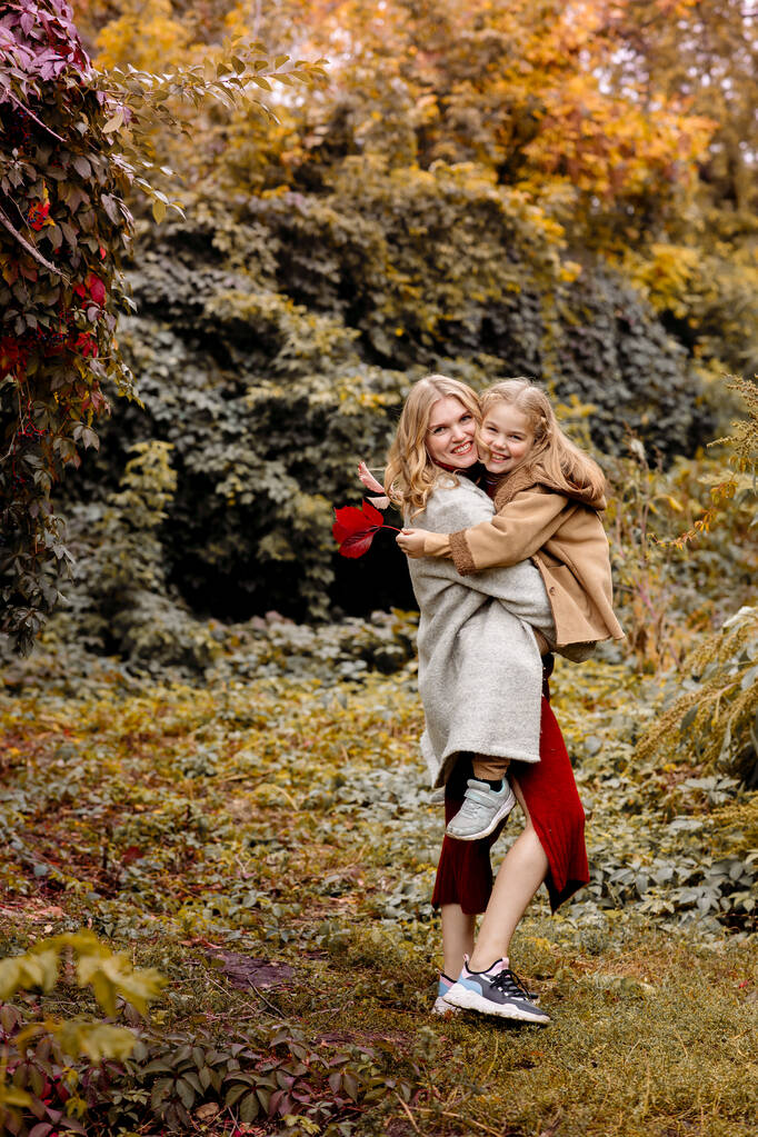Молодая взрослая блондинка мать с 7-летней дочерью на прогулке в осеннем парке обнимаются и смеются - Фото, изображение