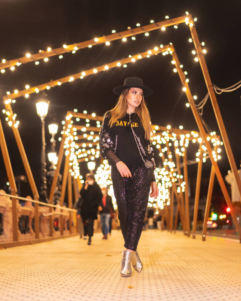 Kerstverlichting in de stad, Kaukasisch blond meisje gekleed in het zwart en met lovertjes op een verlichte brug, winterse levensstijl - Foto, afbeelding