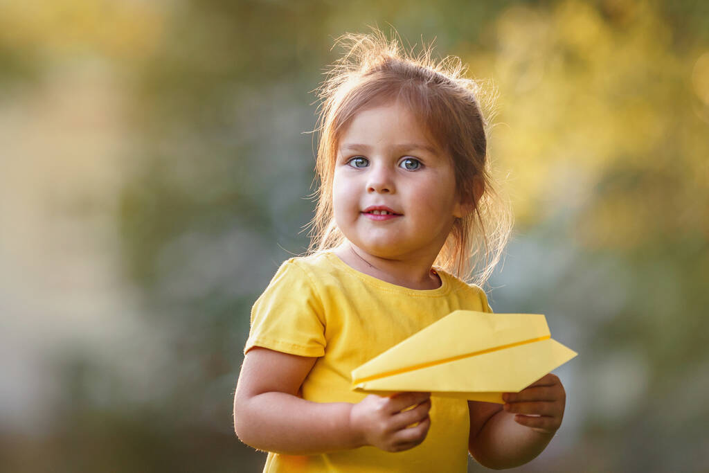 Портрет милой маленькой девочки в желтой футболке стоит с бумажным самолетом в руках. ребенок играет роль пилота, запускающего бумажный самолет в воздух. Малыш. Активная детская игра на свежем воздухе. - Фото, изображение