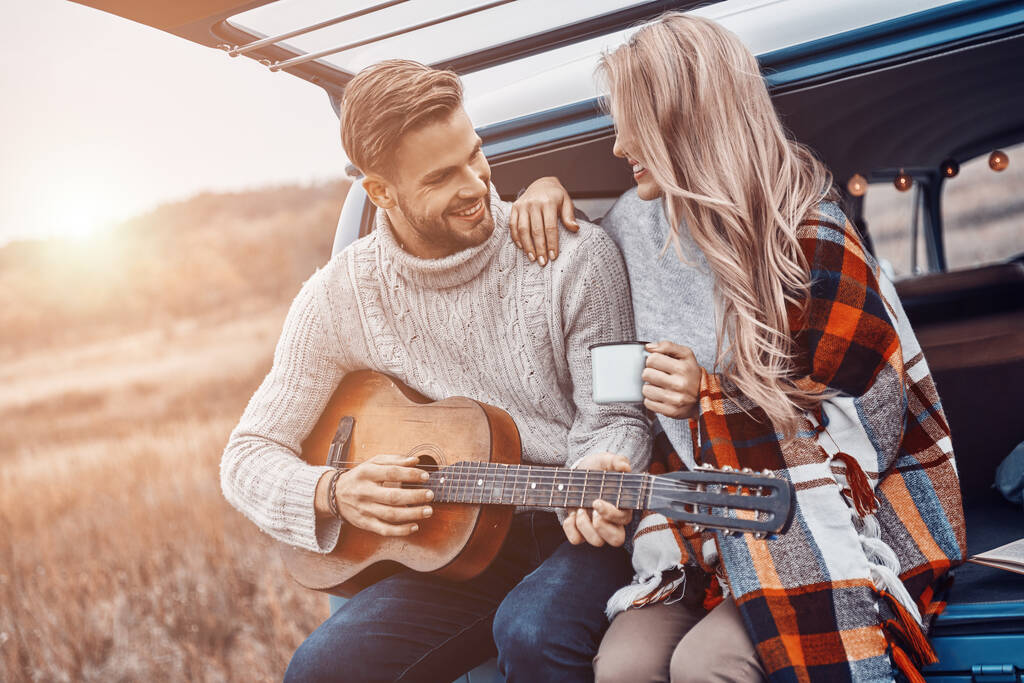 Schöner junger Mann spielt Gitarre für seine Freundin, während beide im Kofferraum sitzen - Foto, Bild
